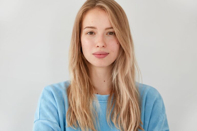Close up shot of pleasant looking serious blonde Caucasian female with long hair, wears blue casual sweater, looks directly into camera, isolated over white background
